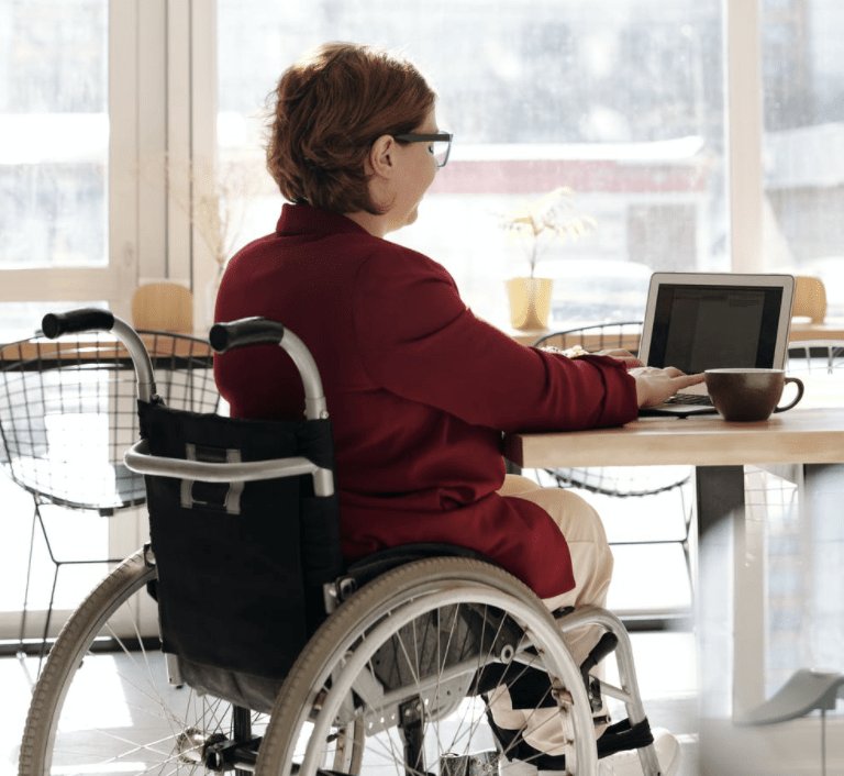 A lady in a red jacket sits in a wheelchair inside at a laptop computer