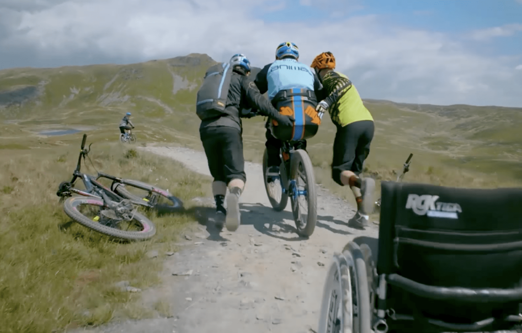 A man in a blue jacket and helmet is helped on his bike by two people