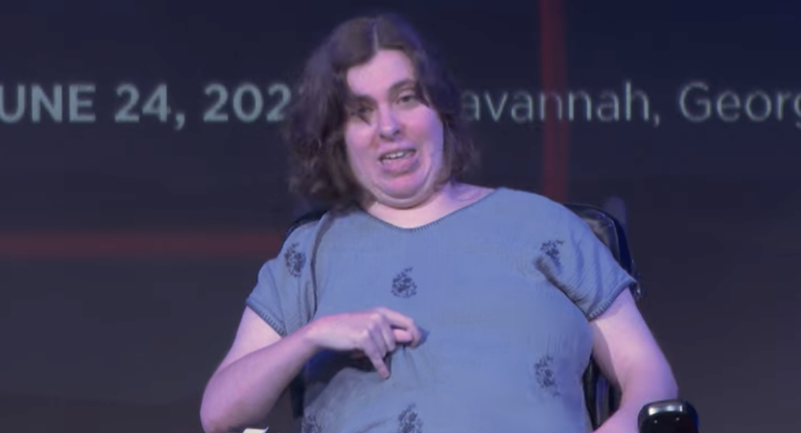 A lady in a blue top talks to an audience on stage in a wheelchair