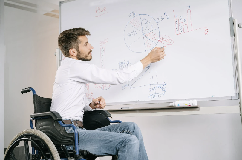 A man in a white short and jeans writes on a whiteboard as a disabled worker in a wheelchair